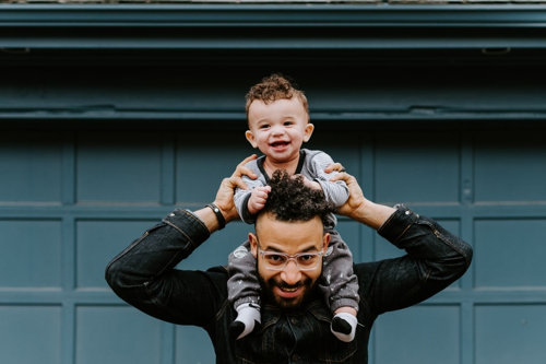 Photograph of a father with his toddler sitting on his shoulders, his arms steading the young child and both smiling.
