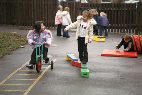 two young girls talking in a playground and smiling, one is cycling on a tricycle, the other balancing on a beam